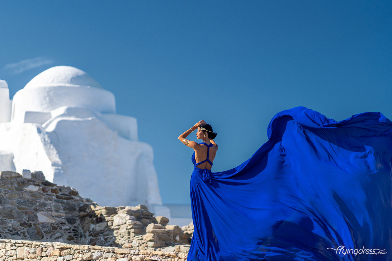 Woman in a royal blue flying dress showcasing the open back design, with the dress flowing dramatically in the wind against white-washed Mykonos architecture and a deep blue sky.